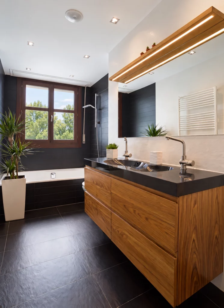 Fotografía de interiorismo de un baño moderno con mueble suspendido de madera, encimera negra de doble seno, espejo con iluminación LED lineal y bañera junto a la ventana.